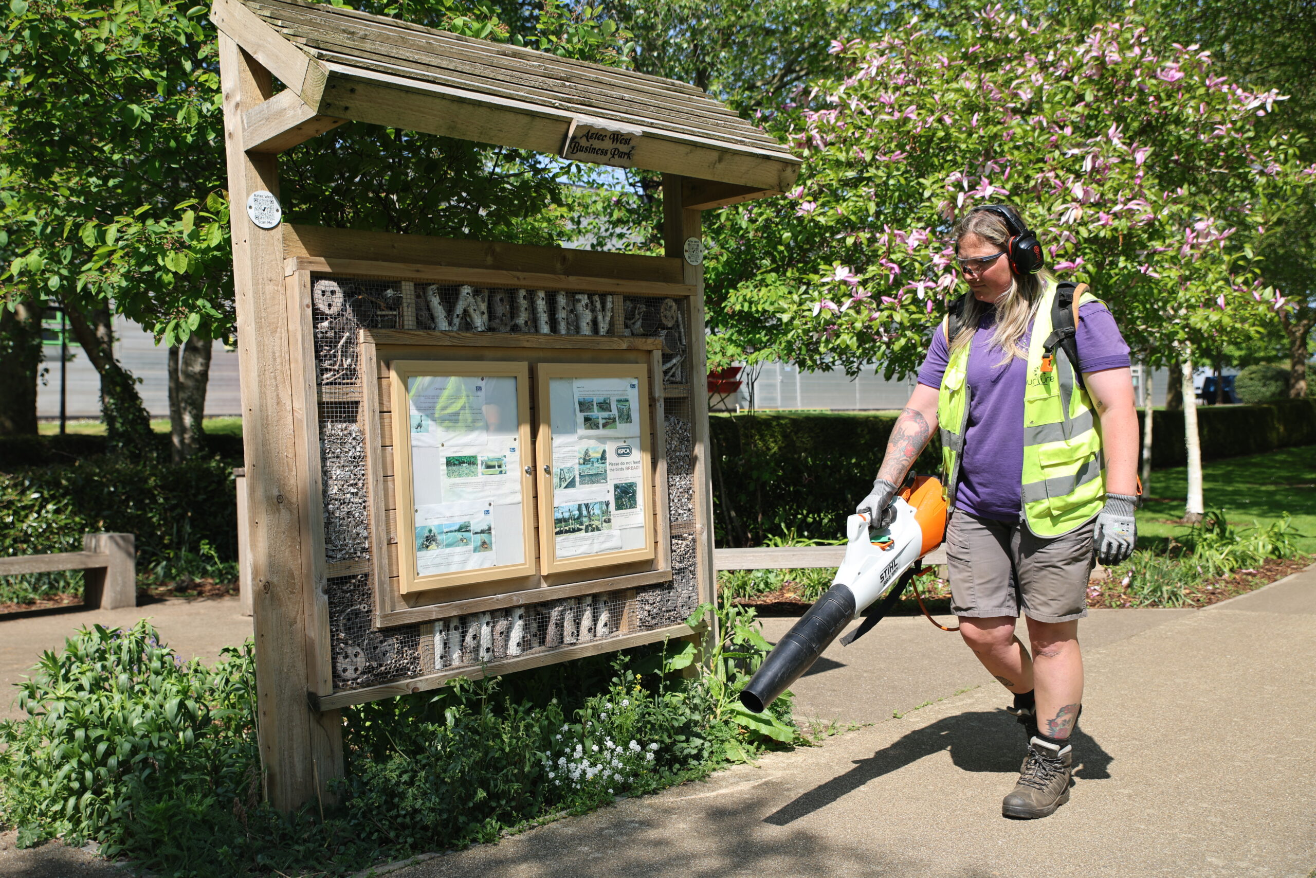 Female grounds maintenance operative working with battery operated equipment in front of a bespoke insect habitat sign